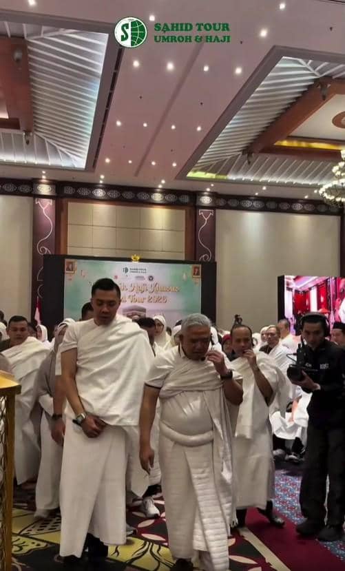 Sekelompok jamaah pria mengenakan pakaian ihram putih sedang mengikuti manasik haji di sebuah aula dengan dekorasi bertema haji.
