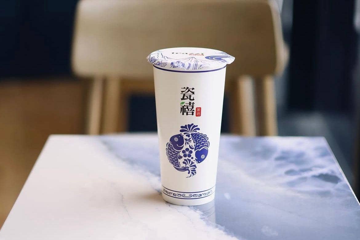Gelas minuman teh Teazzi berdesain ikan biru dengan tulisan Mandarin di atas meja marmer di dalam ruangan.