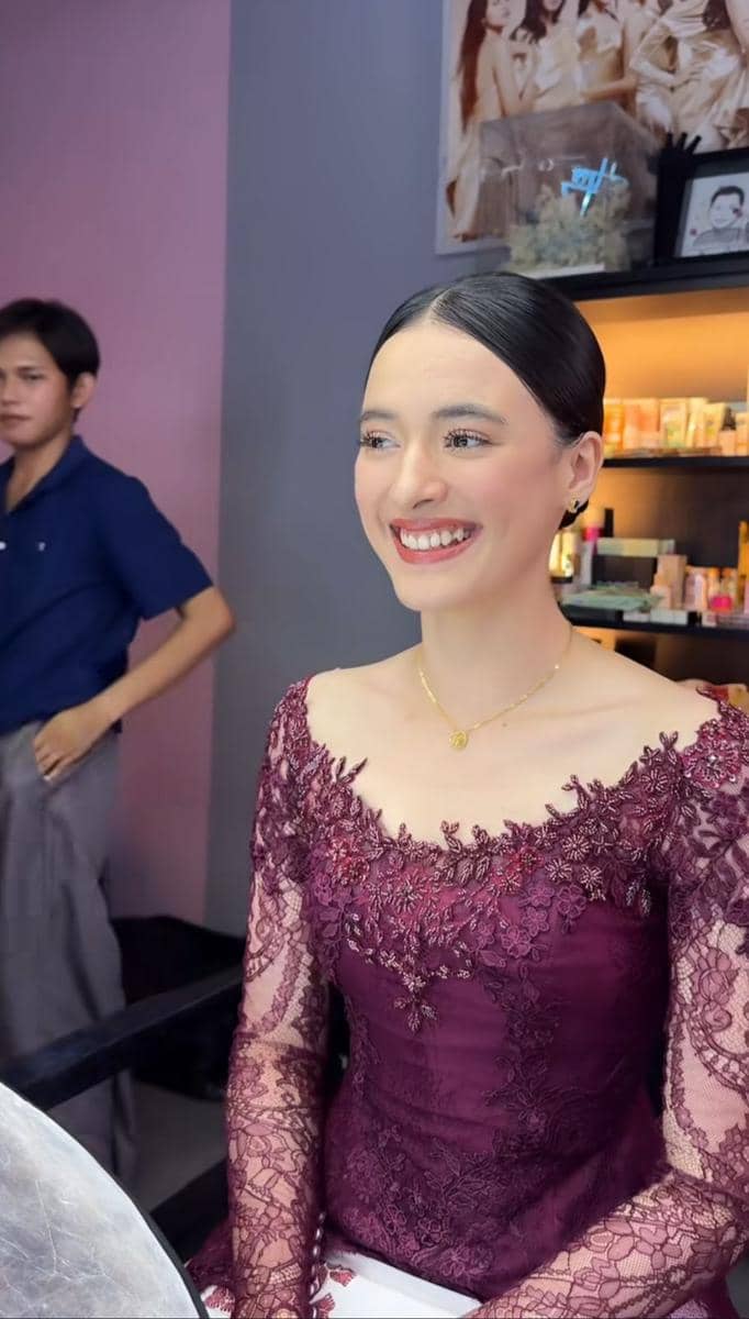 Seorang perempuan tersenyum mengenakan kebaya ungu marun berhias renda saat bersiap untuk acara wisuda di ruang rias.
