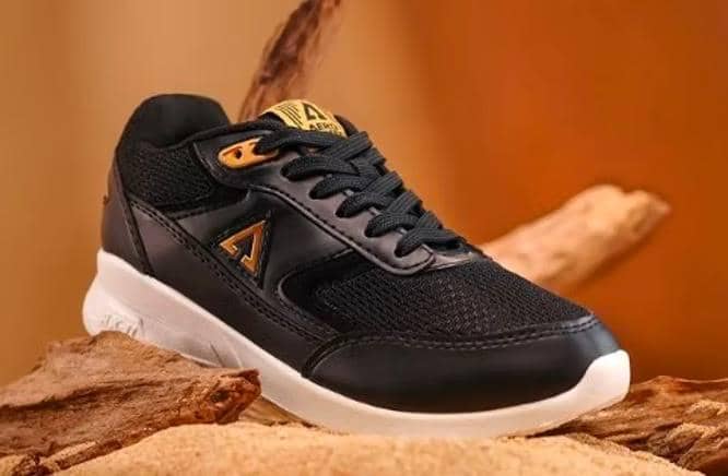 Sepatu sneakers hitam Aerostreet dengan sol putih dan detail logo emas, diletakkan di atas permukaan kayu dengan latar cokelat hangat.