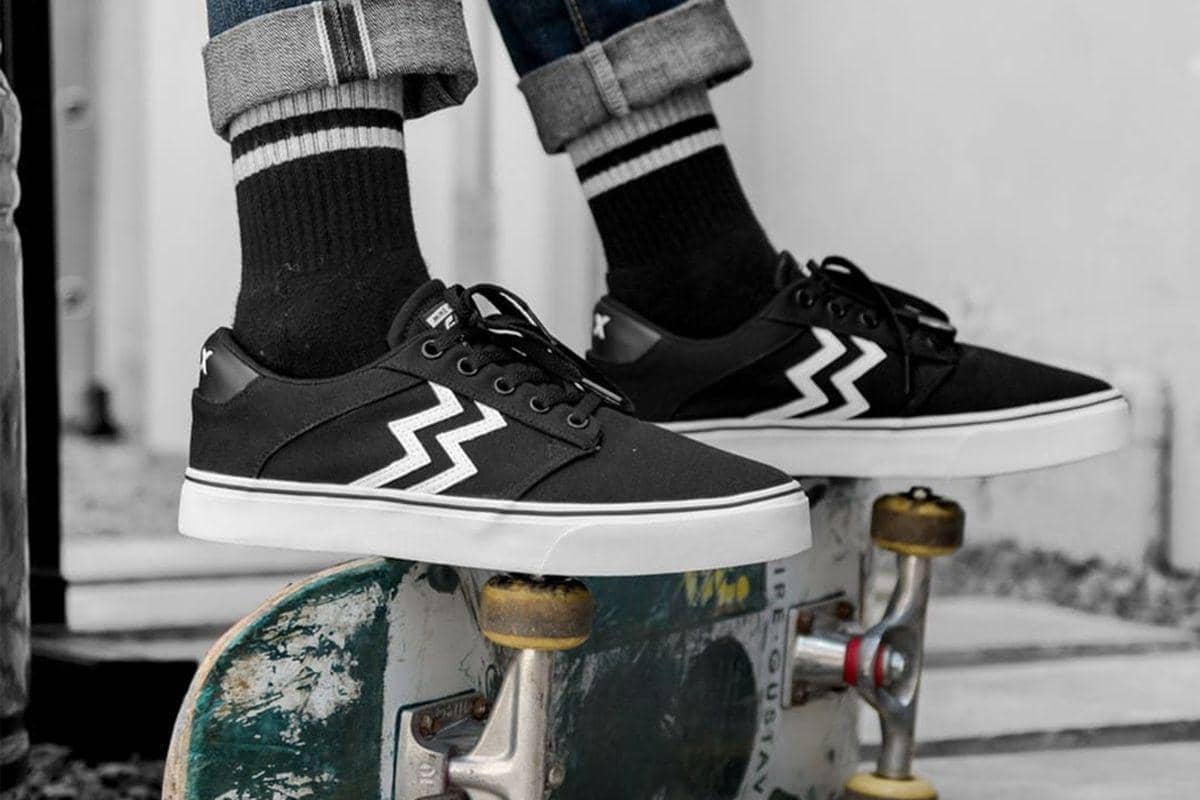 Sepasang sepatu skate hitam dengan sol putih dan motif garis zigzag putih dikenakan oleh seseorang yang berdiri di atas papan skateboard.