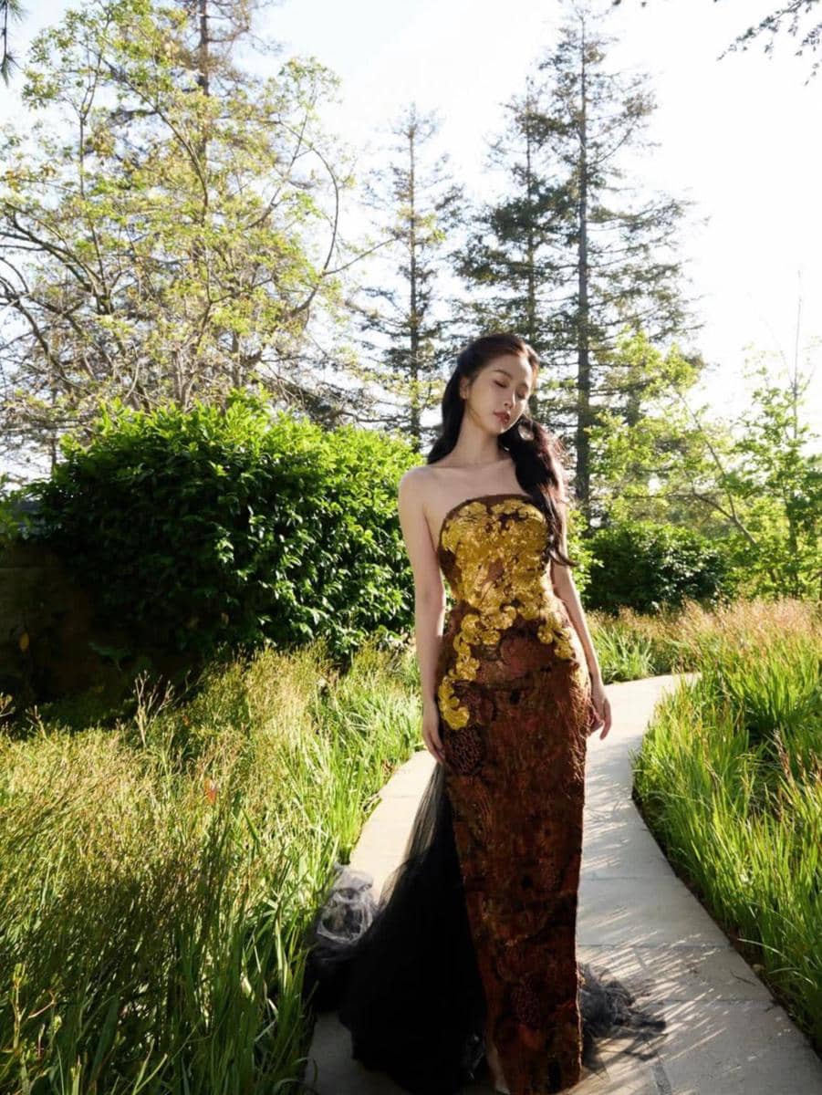Seorang wanita mengenakan gaun panjang berwarna emas dan hitam dengan detail bunga berjalan di taman dengan latar pepohonan hijau.