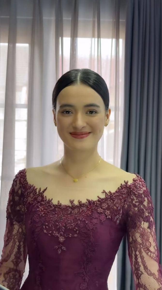Seorang perempuan mengenakan kebaya ungu marun dengan detail renda halus, rambut disanggul rapi, dan tersenyum di depan tirai.
