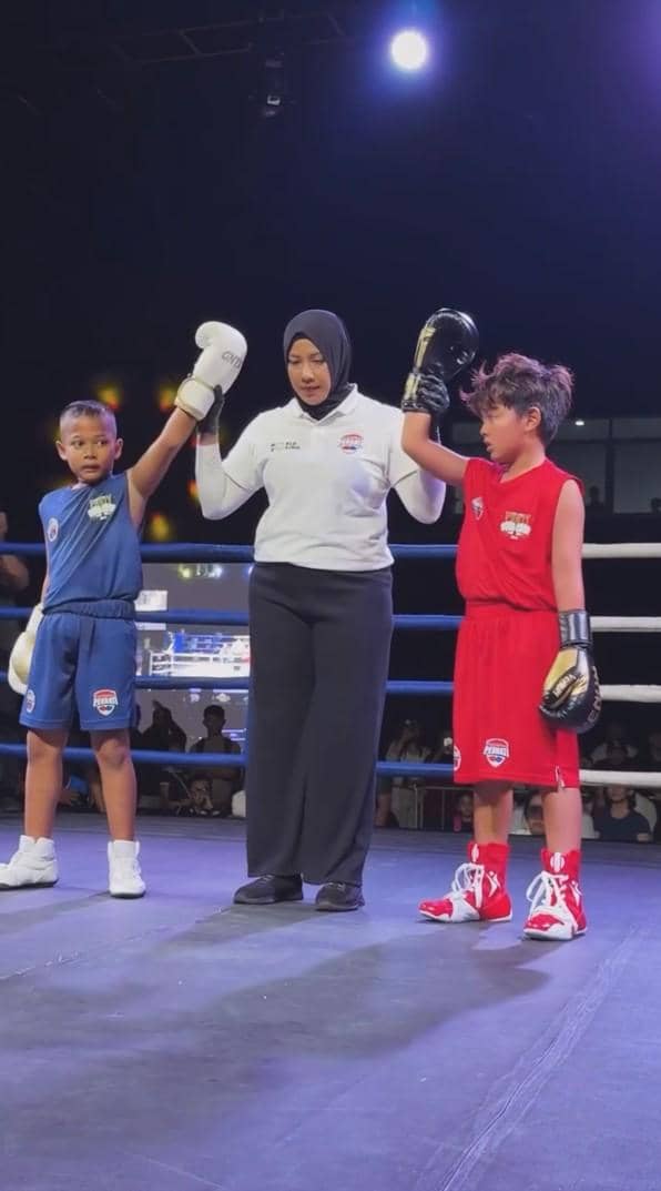 Dua anak mengenakan perlengkapan tinju biru dan merah berdiri di atas ring bersama wasit perempuan yang mengangkat tangan mereka.