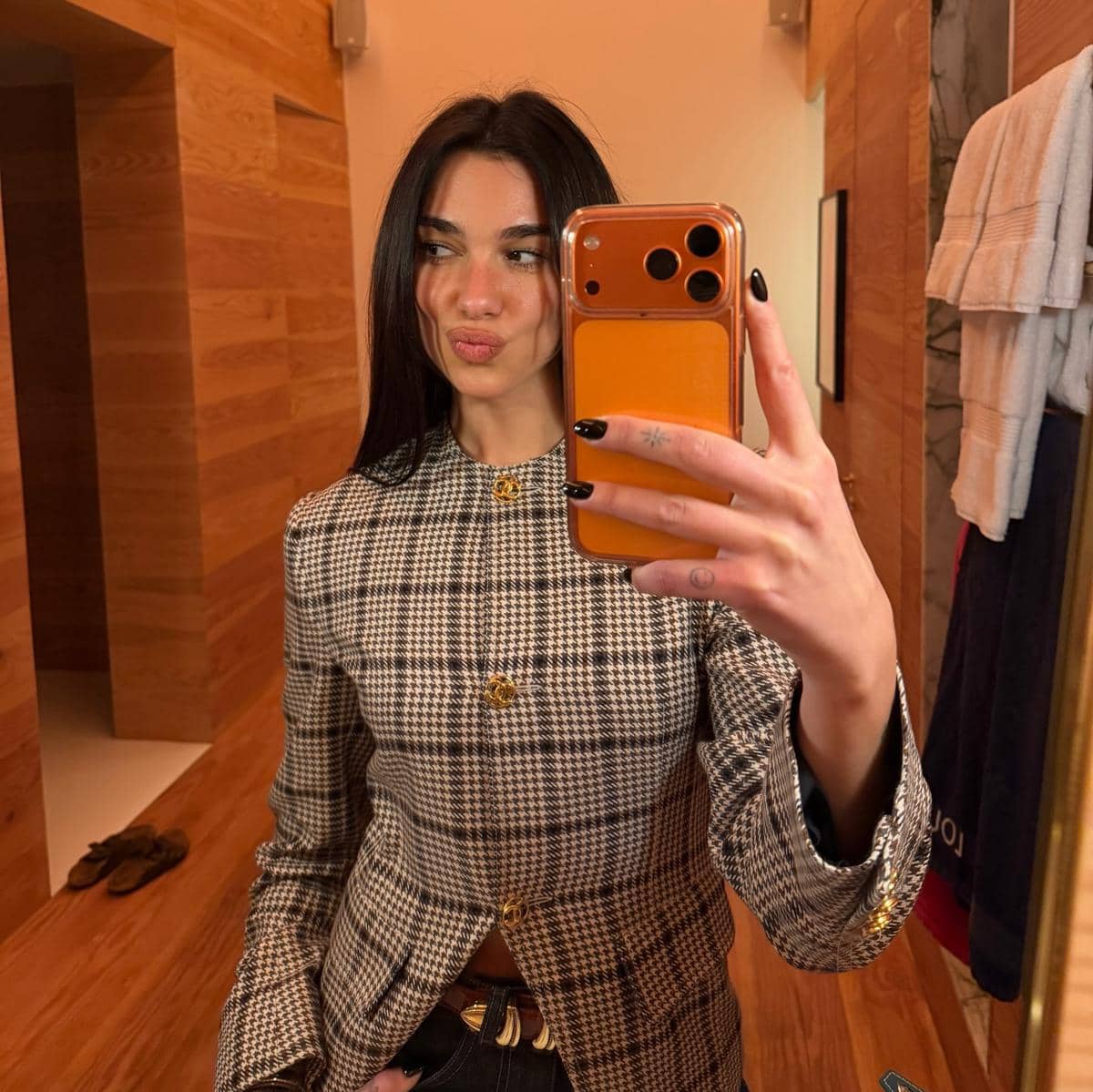 Seorang perempuan berambut hitam panjang berpose selfie di depan cermin dengan ponsel berwarna oranye, mengenakan blazer motif kotak-kotak.