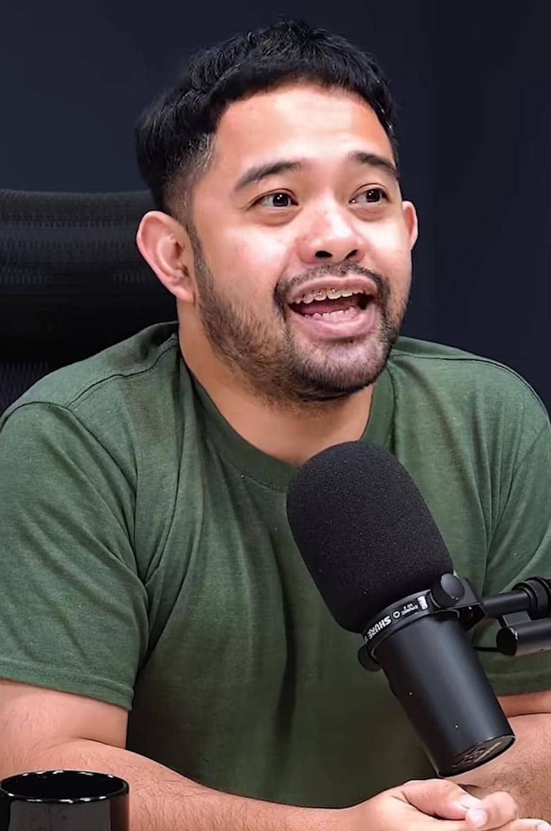 Seorang pria berjanggut mengenakan kaus hijau sedang berbicara di depan mikrofon hitam di studio dengan latar belakang gelap.