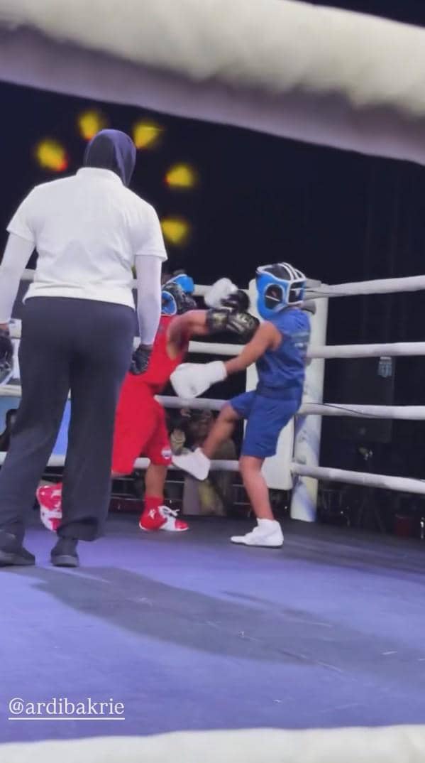 Dua anak mengenakan perlengkapan tinju lengkap sedang bertanding di atas ring dengan wasit mengawasi dari dekat.