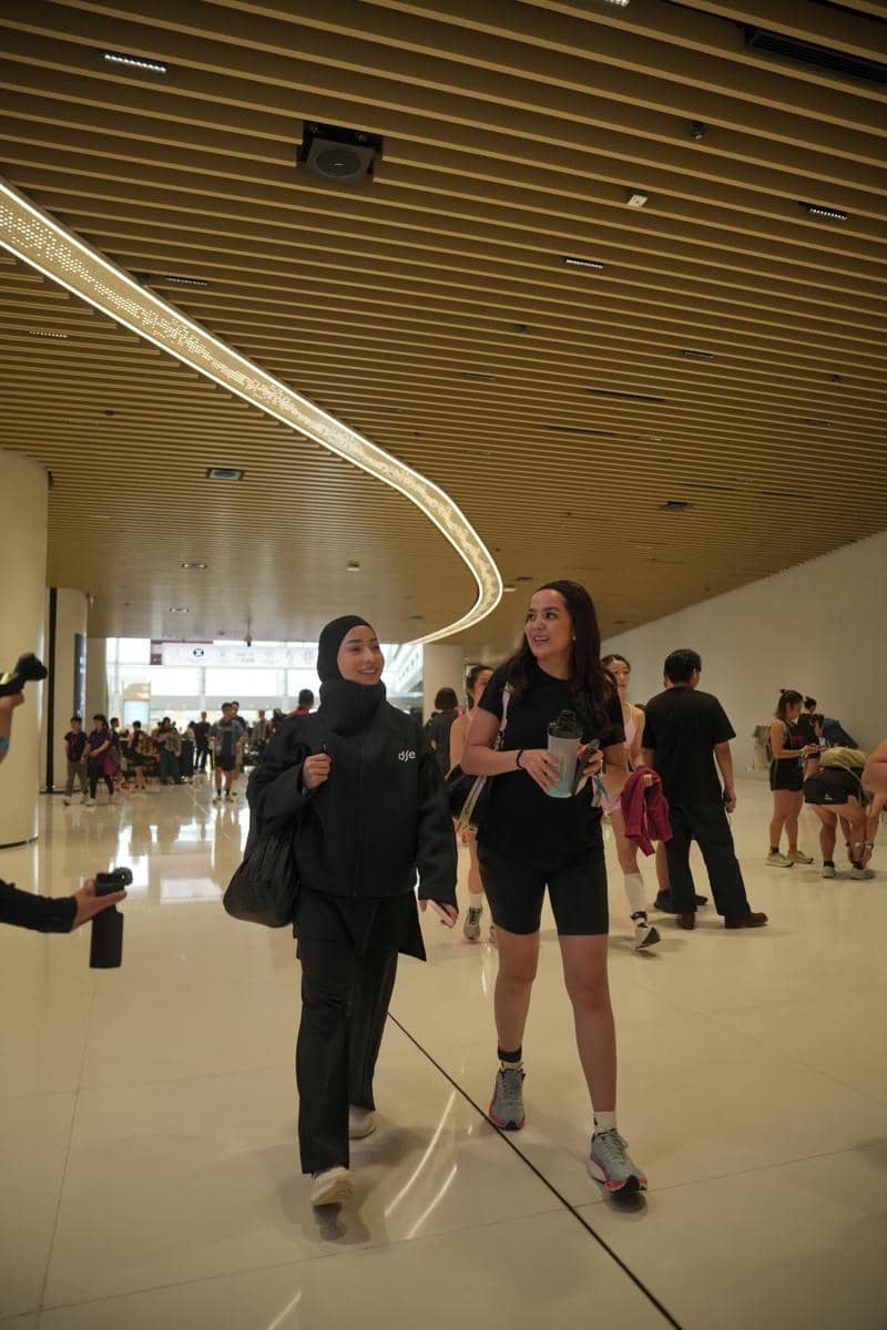 Dua perempuan berjalan bersama di area dalam gedung modern dengan langit-langit kayu dan pencahayaan melengkung di Hong Kong.