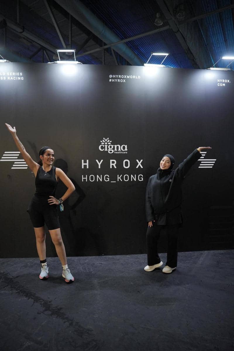 Dua perempuan berpose di depan latar bertuliskan Hyrox Hong Kong dengan pencahayaan terang di area dalam ruangan.