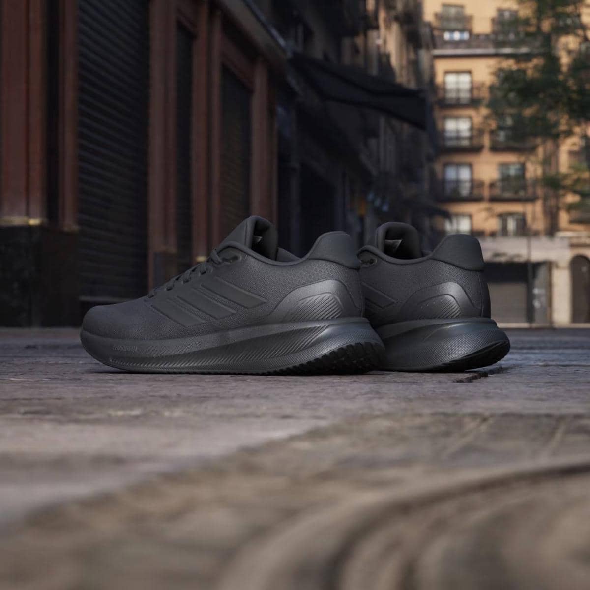 Sepasang sepatu lari Adidas Runfalcon 5 warna hitam diletakkan di jalan kota dengan latar gedung bertingkat dan suasana tenang.