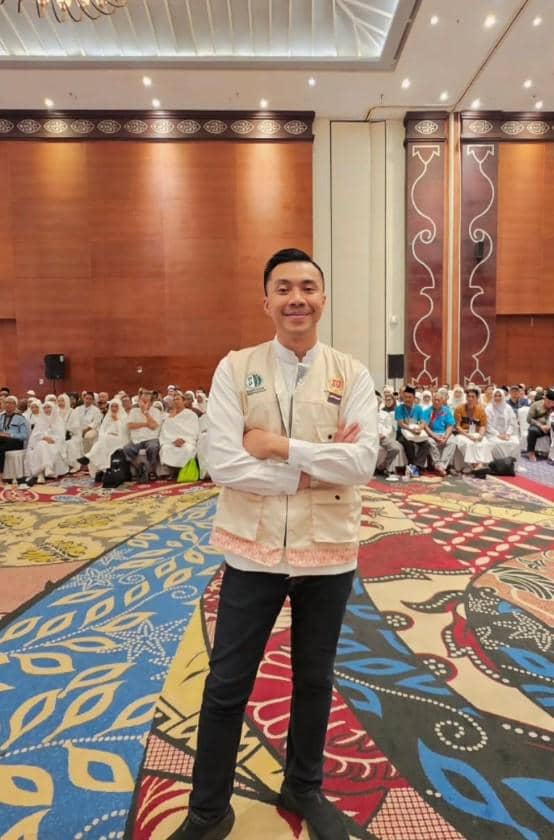Seorang pria berdiri dengan tangan terlipat di aula besar, di belakangnya terdapat jamaah haji yang mengenakan pakaian putih.