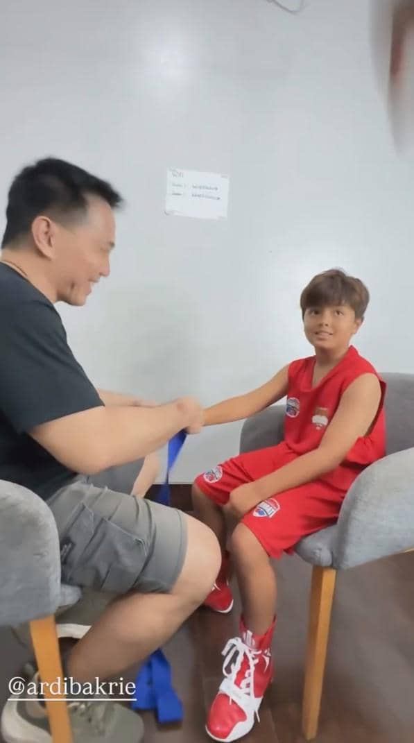 Seorang anak mengenakan seragam tinju merah duduk di kursi sambil dibantu pria dewasa membalut tangannya sebelum pertandingan.