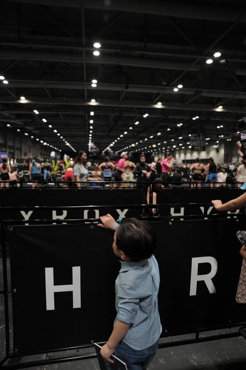 Seorang anak kecil berdiri di dekat pagar bertuliskan HYROX di dalam arena olahraga indoor yang ramai dengan peserta dan penonton.