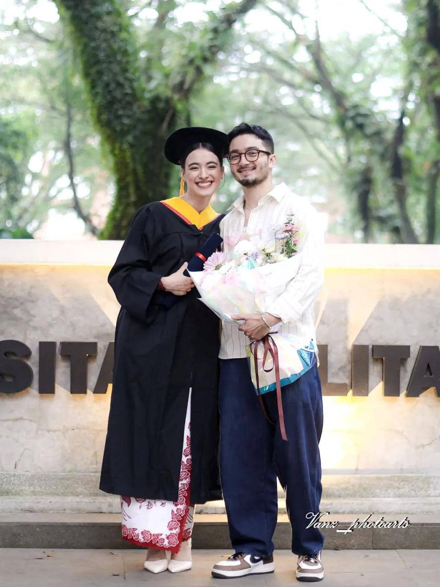 Seorang wisudawati mengenakan toga hitam dan kebaya merah putih berpose bersama seorang pria yang membawa buket bunga di depan area kampus.
