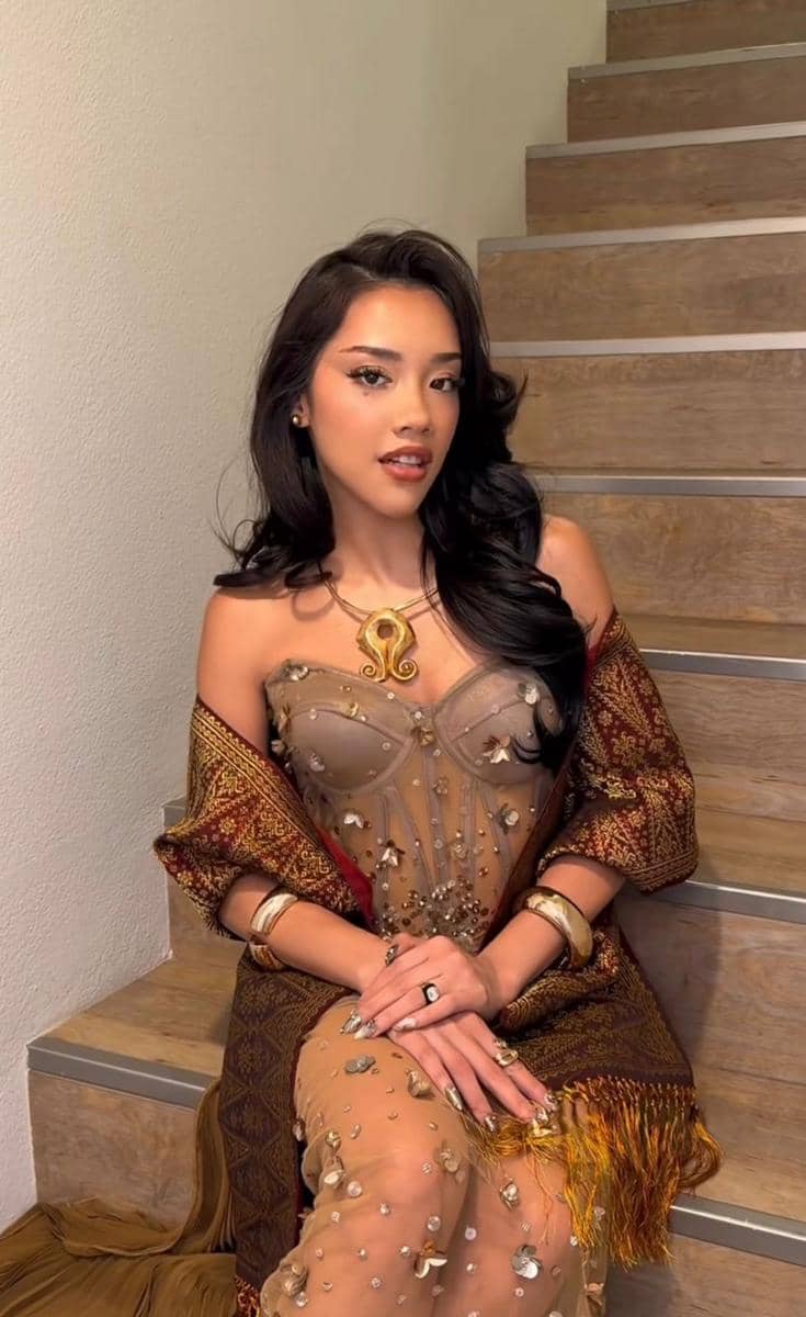 Seorang wanita duduk di tangga mengenakan gaun transparan berhias payet dan kain tradisional bermotif etnik dengan aksesori emas.