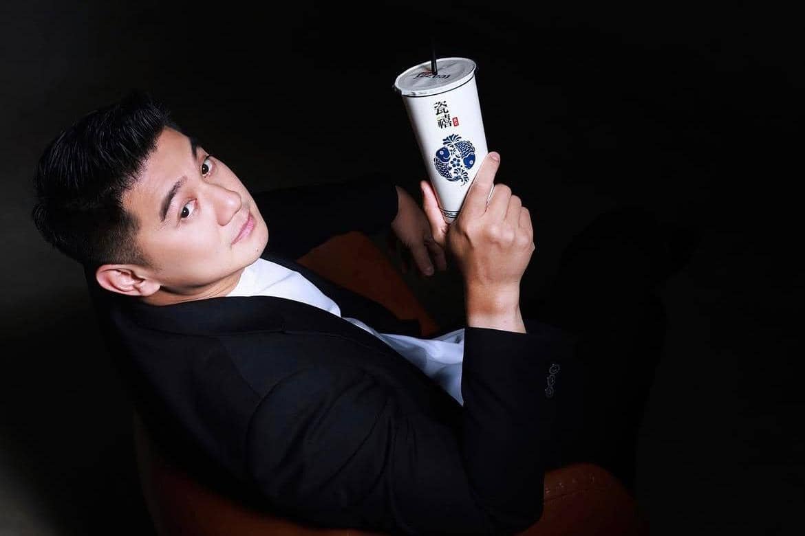 Seorang pria mengenakan jas hitam dan kemeja putih memegang gelas minuman bermerek Teazzi dengan latar belakang gelap.
