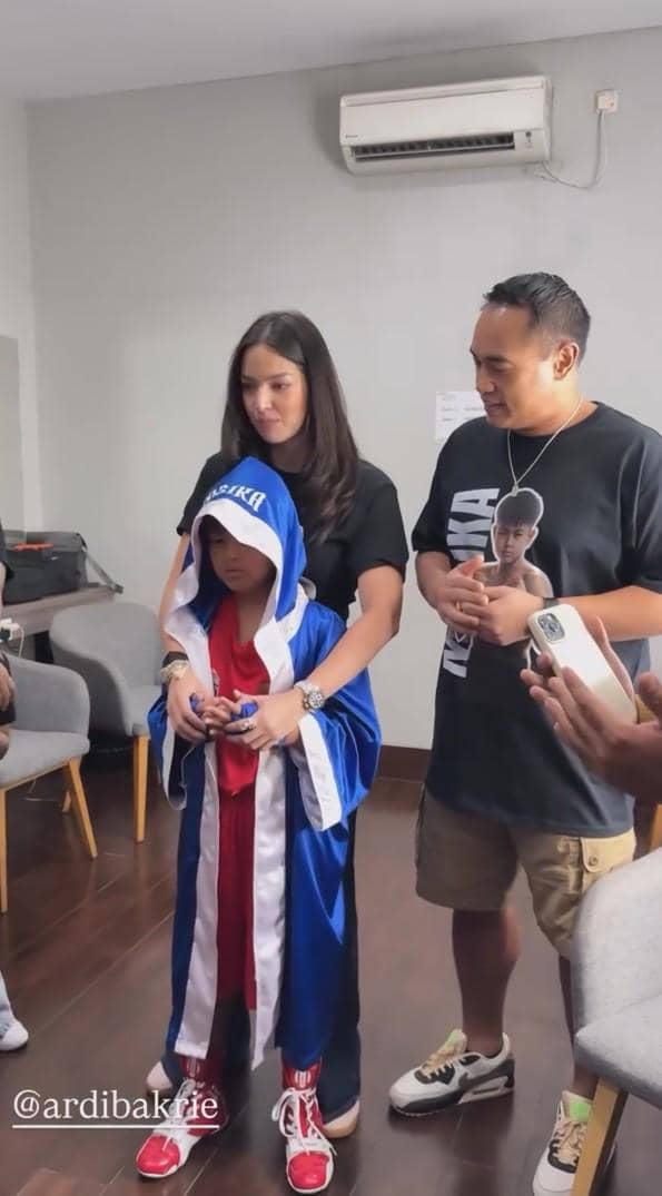 Seorang anak mengenakan jubah tinju biru dan merah berdiri bersama dua orang dewasa di dalam ruangan sebelum pertandingan tinju.