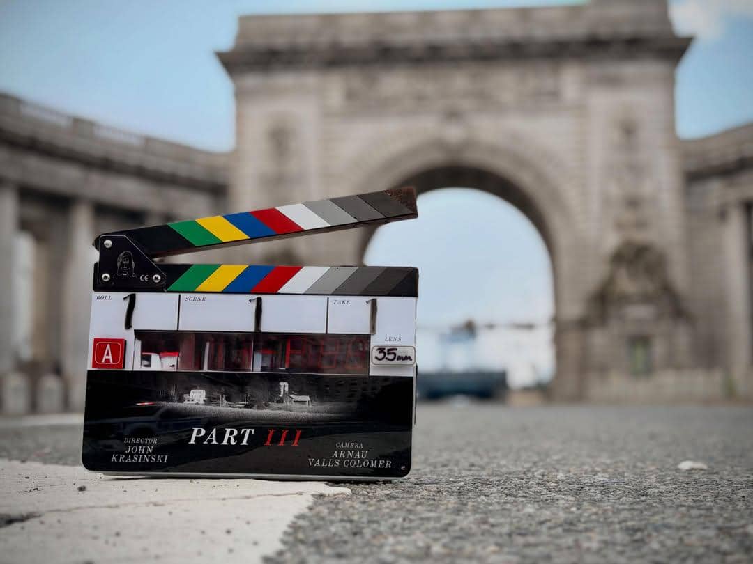 Papan clapperboard bertuliskan A Quiet Place Part 3 berdiri di jalan dengan latar lengkungan batu besar di kejauhan.