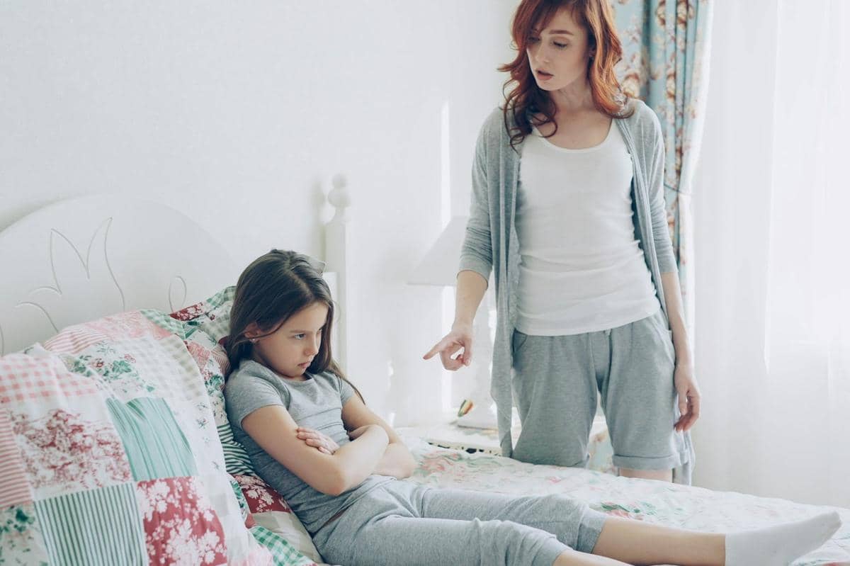 Seorang ibu berbicara dengan anak perempuannya yang tampak marah dan menyilangkan tangan di atas tempat tidur di kamar bernuansa lembut.