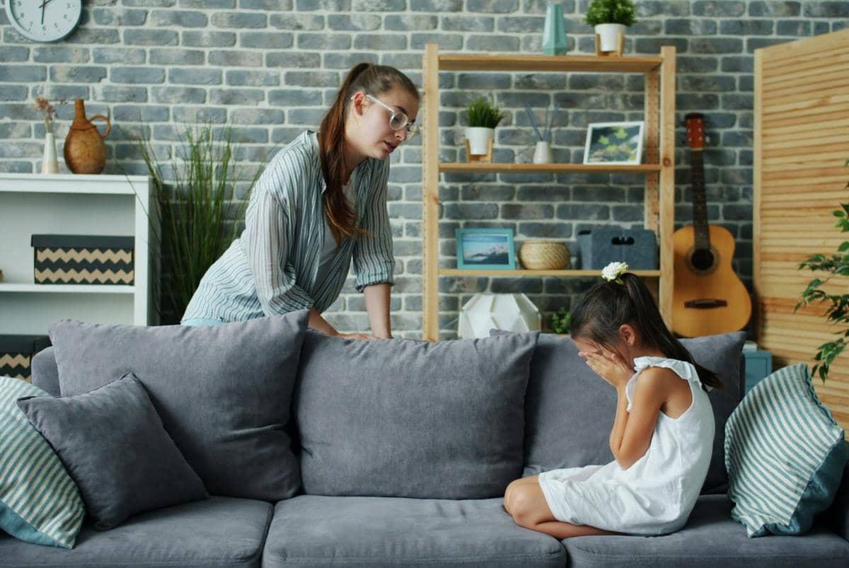 Seorang ibu membujuk anak perempuannya yang sedang menangis di ruang tamu dengan sofa abu-abu dan dinding bata abu-abu.