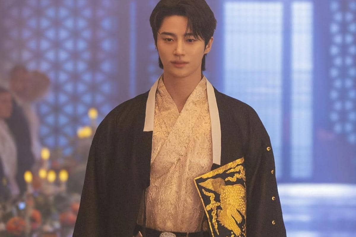 Seorang aktor mengenakan hanbok modern berwarna hitam dan emas dengan latar ruangan bercahaya lembut dalam adegan drama Korea Perfect Crown.