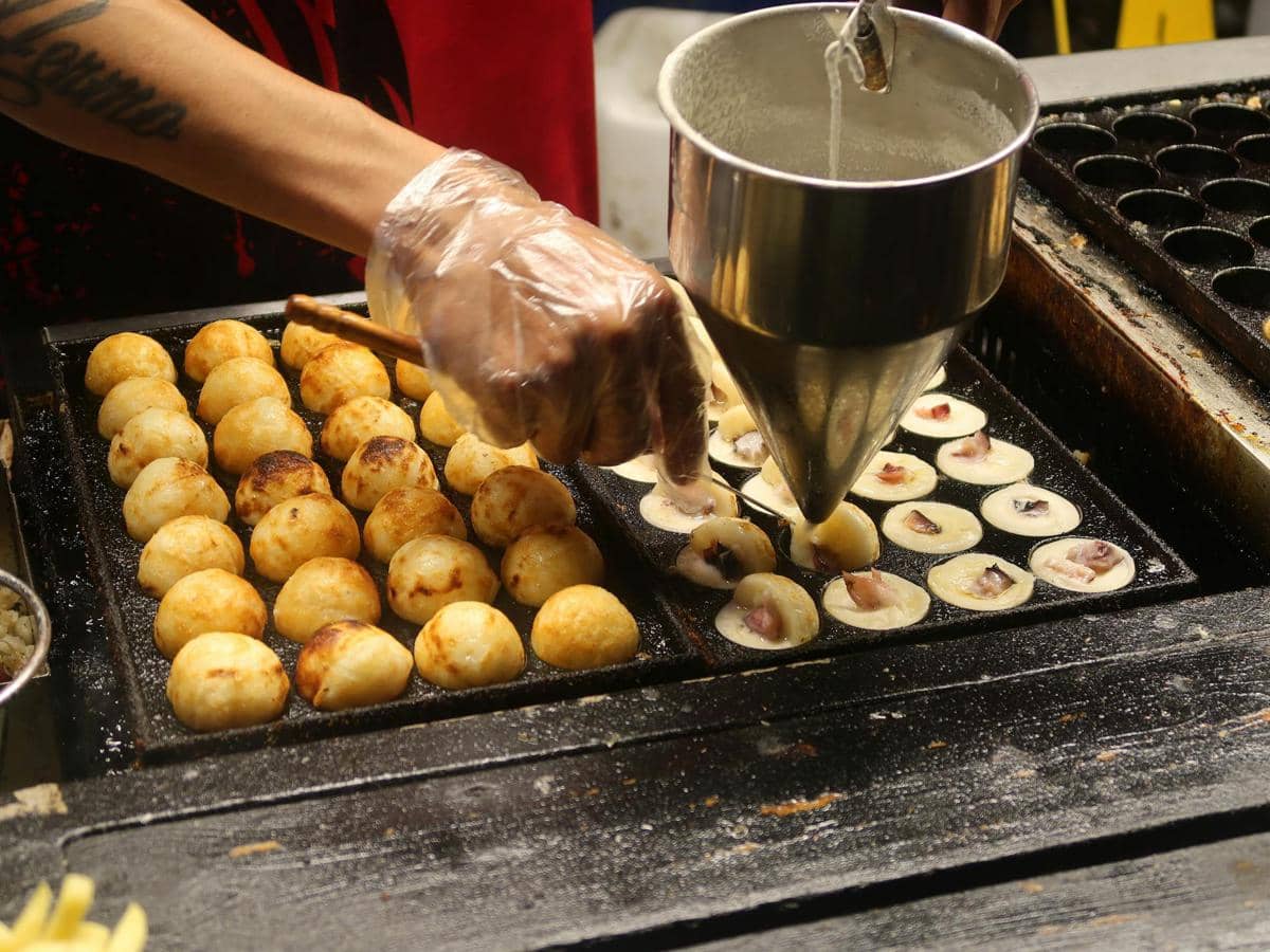 Seorang penjual menuang adonan ke dalam cetakan bulat untuk membuat takoyaki di atas panggangan besi panas.