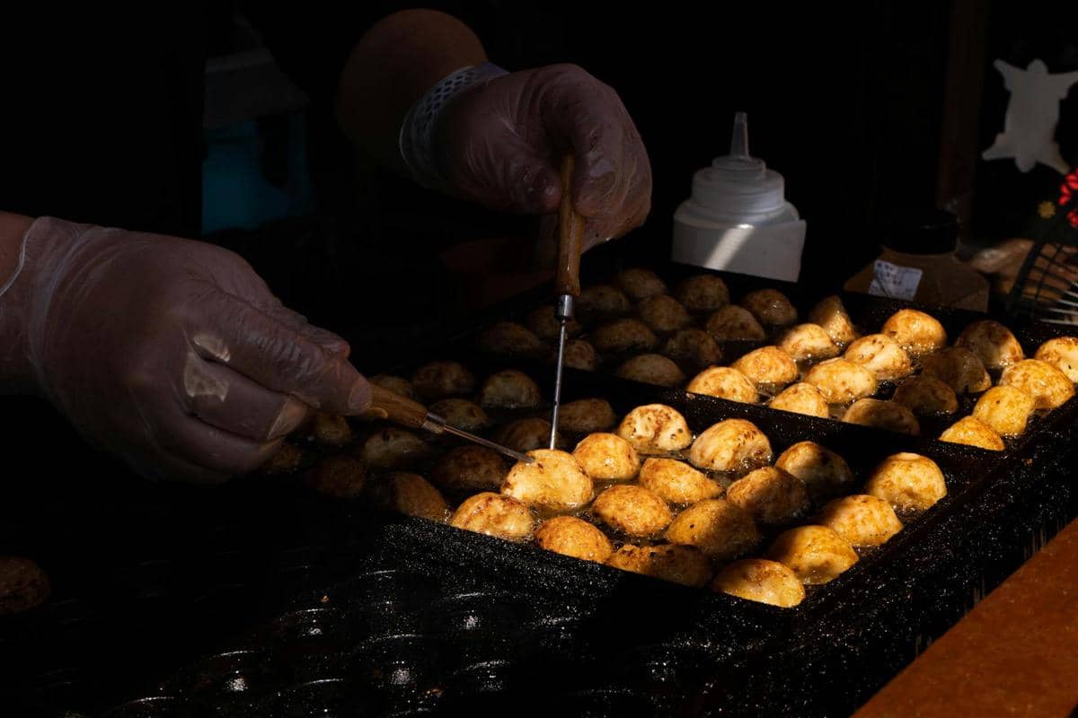Tangan mengenakan sarung tangan sedang membalik adonan takoyaki di atas cetakan besi panas menggunakan tusuk logam.
