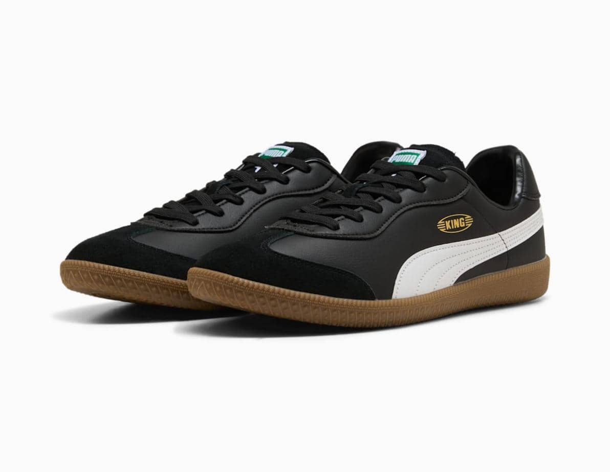 Sepatu futsal King 21 IT berwarna hitam dengan garis putih di sisi dan sol karet cokelat, menampilkan desain klasik dan elegan.