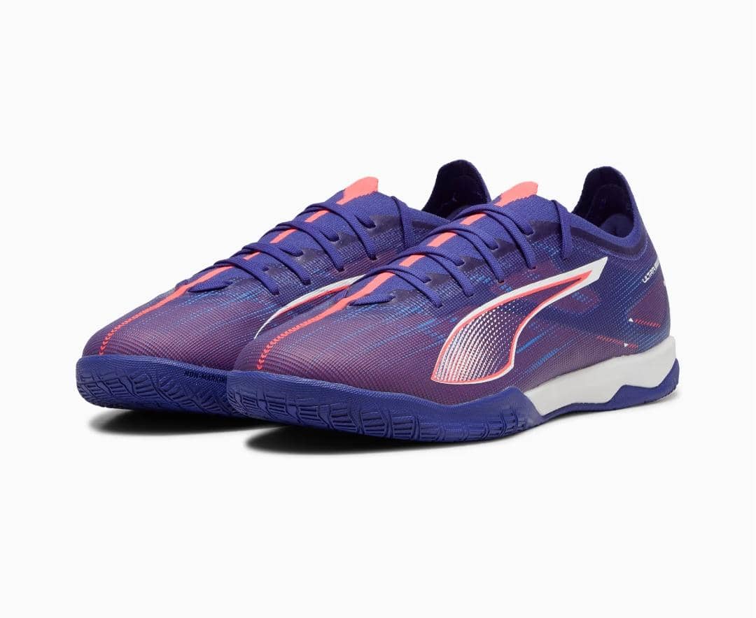 Sepasang sepatu futsal Puma ULTRA 5 MATCH IT berwarna ungu dengan aksen merah muda dan putih, dirancang untuk permainan indoor.