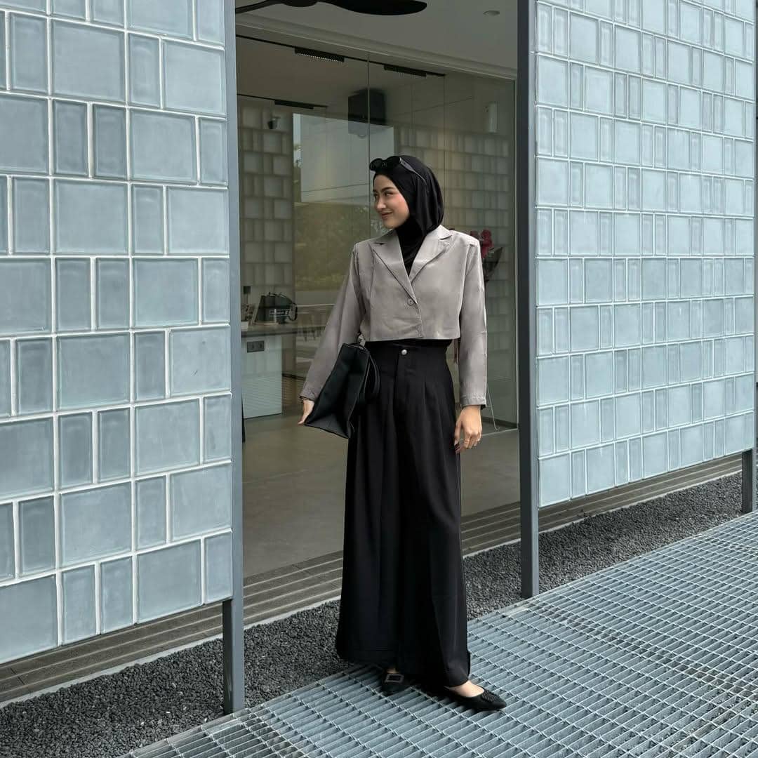 Seorang wanita berhijab mengenakan blazer abu-abu pendek dan celana hitam lebar sambil memegang tas hitam di depan bangunan modern.