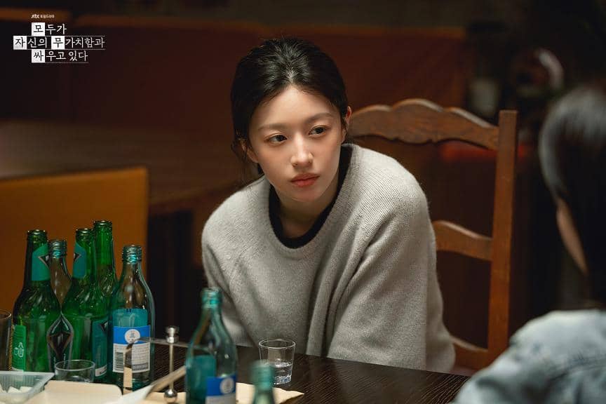 Seorang wanita muda mengenakan sweater abu-abu duduk di meja kayu dengan beberapa botol minuman di depannya dalam adegan drama We Are All Trying Here.