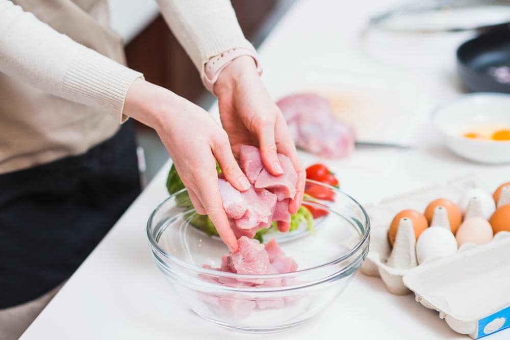 Seseorang memegang potongan daging kambing mentah di atas mangkuk kaca di dapur dengan bahan masakan seperti telur dan sayuran di sekitarnya.