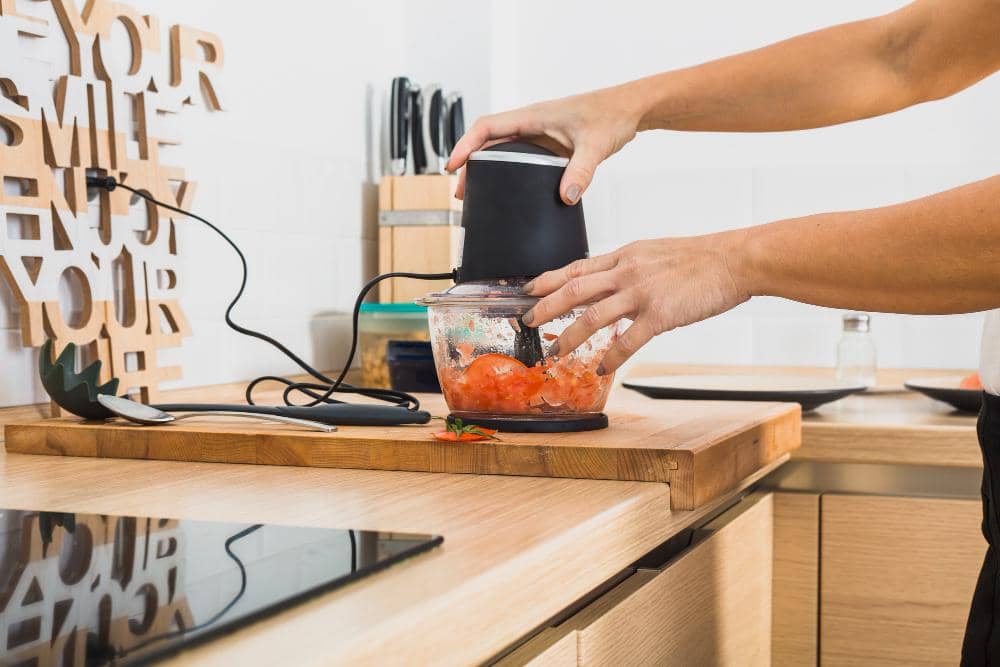 Seseorang menggunakan blender kecil di dapur modern untuk menghaluskan tomat di atas meja kayu dengan peralatan dapur di sekitarnya.