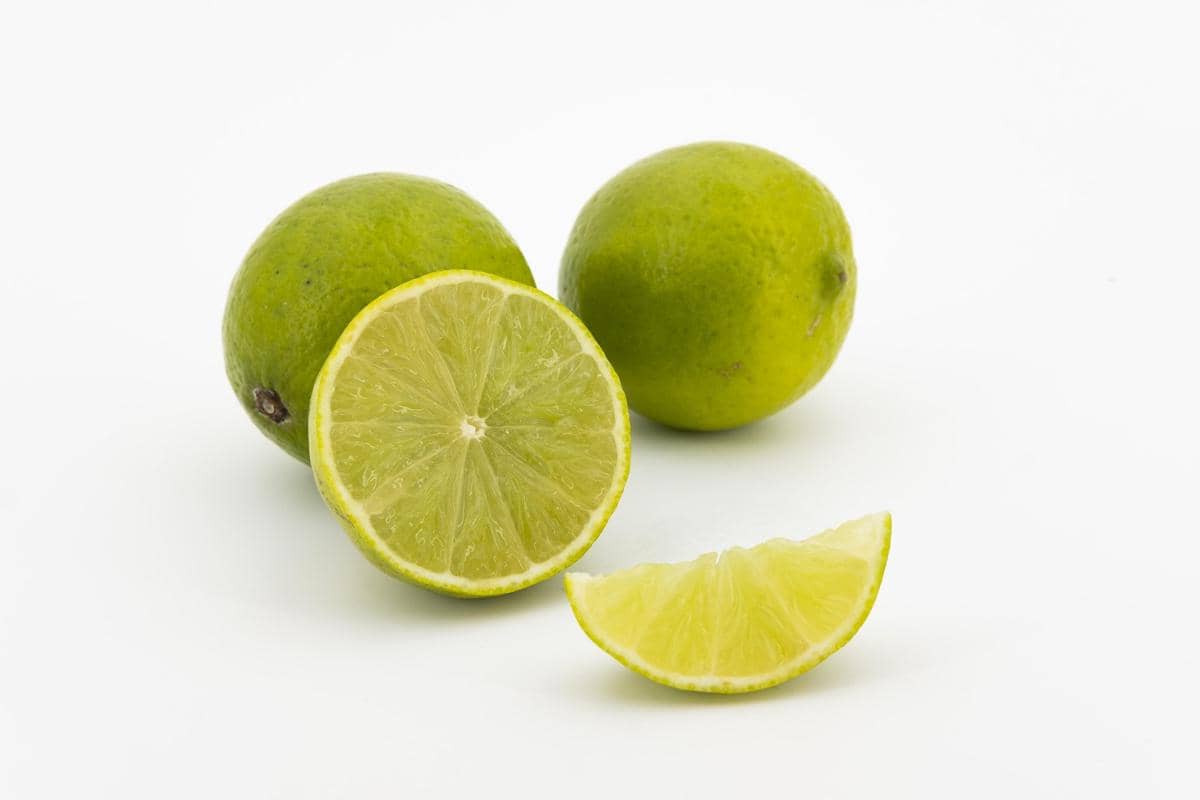 Beberapa buah jeruk nipis segar berwarna hijau dengan satu buah terbelah menampilkan bagian dalam berair di latar putih.