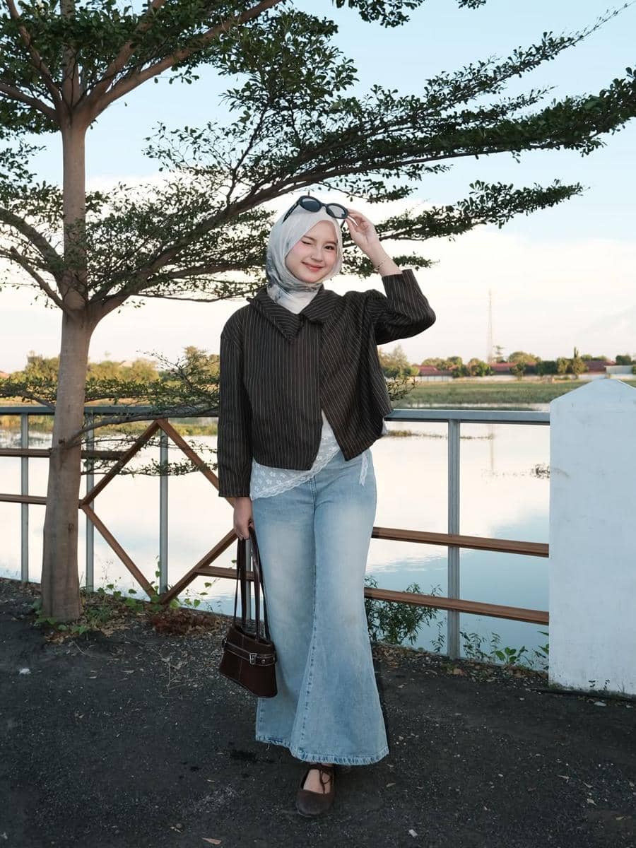 Seorang wanita berhijab mengenakan jaket cokelat bergaris dan celana jeans biru lebar, berpose di tepi danau dengan latar pepohonan.