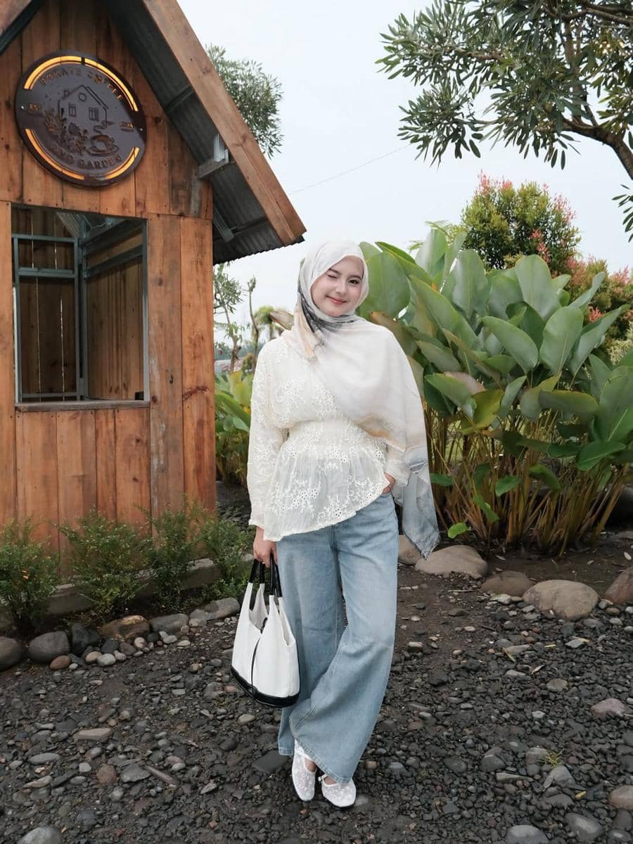 Seorang wanita berhijab mengenakan blus renda putih dan celana jeans biru, berpose di depan bangunan kayu dengan latar taman hijau.