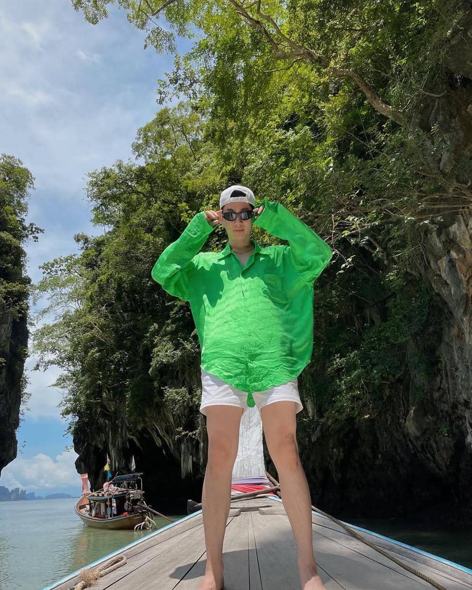 Seorang pria mengenakan kemeja hijau cerah dan celana pendek putih berdiri di atas perahu di antara tebing dan pepohonan tropis.