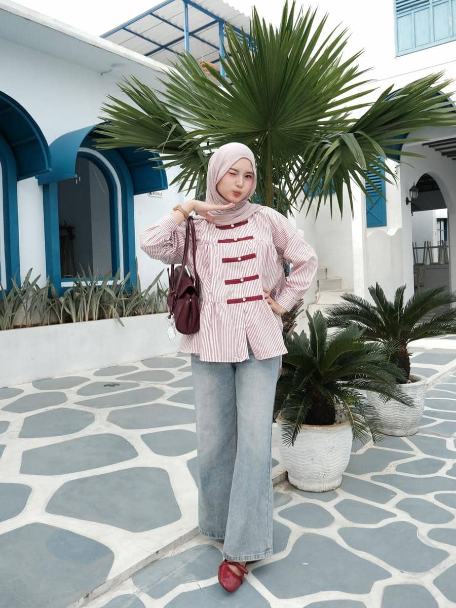 Seorang wanita berhijab mengenakan kemeja bergaris merah muda dan celana jeans biru lebar berpose di area outdoor bergaya Mediterania.