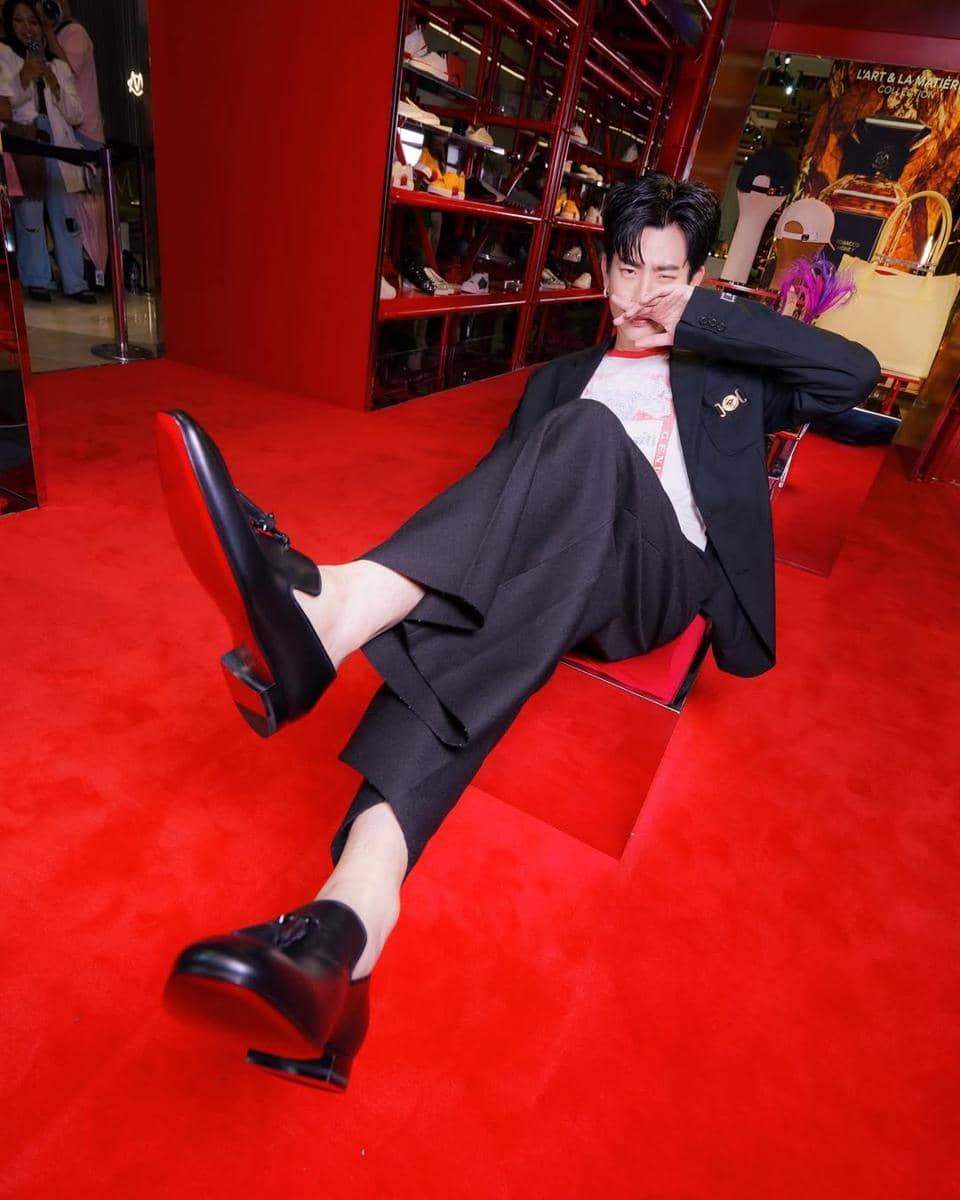 Seorang pria bergaya formal duduk santai di lantai merah dengan setelan jas hitam, celana panjang, dan sepatu kulit hitam elegan.