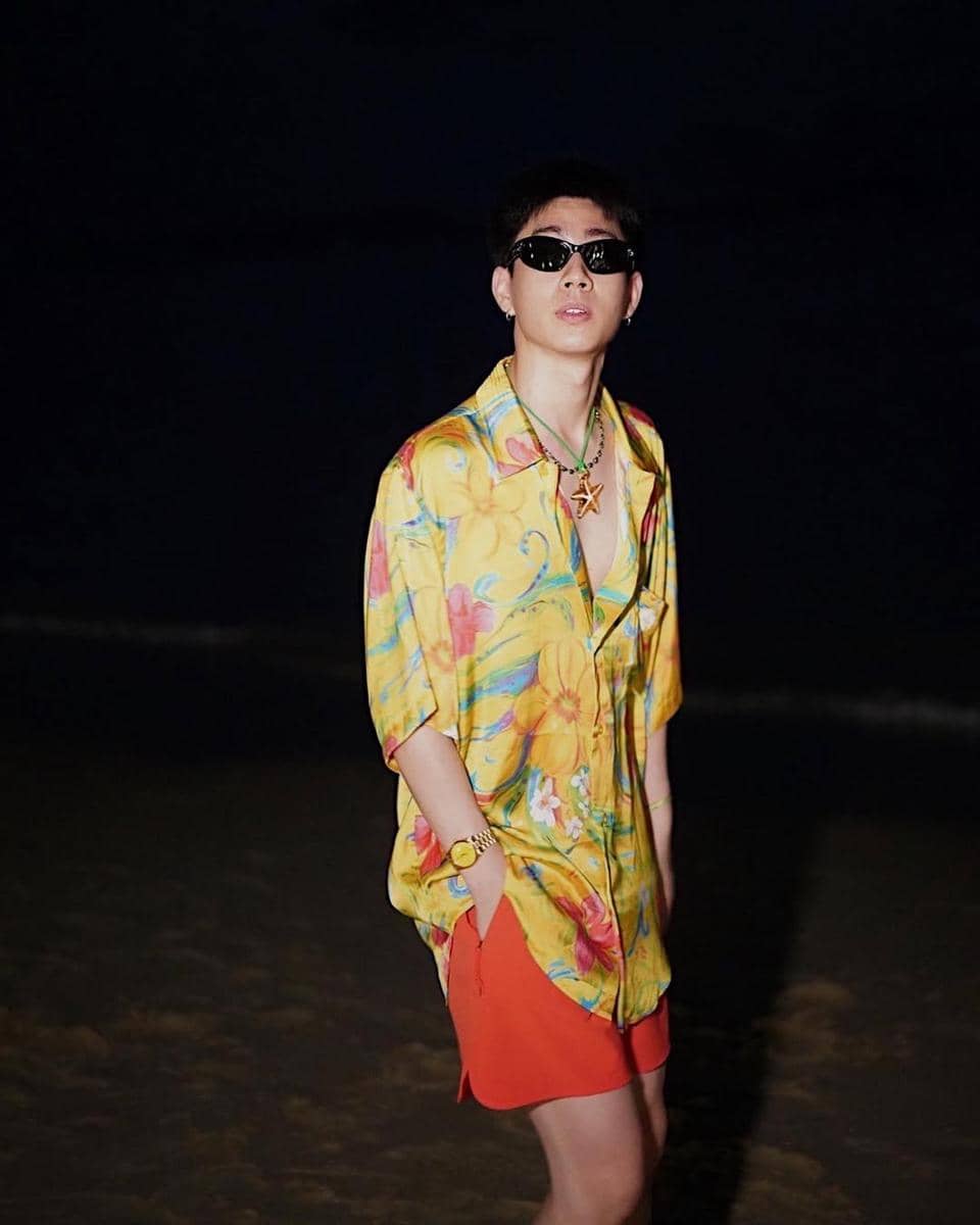 Pria mengenakan kemeja motif bunga berwarna kuning cerah dan celana pendek merah, memakai kacamata hitam di tepi pantai malam hari.