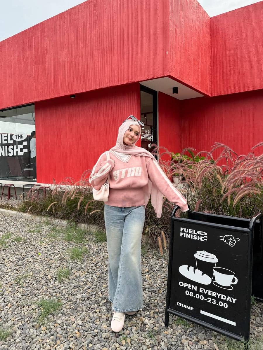 Seorang wanita berhijab mengenakan sweater pink dan celana jeans biru berpose di depan kafe berdinding merah dengan papan informasi jam buka.