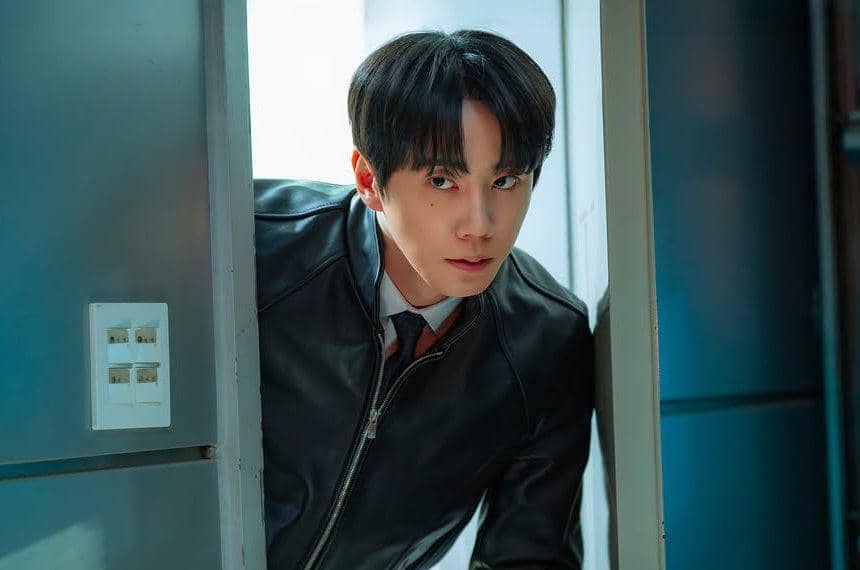 Seorang pria mengenakan jaket kulit hitam dan dasi sedang mengintip dari balik pintu dengan ekspresi serius di adegan drama Korea Reborn Rookie.