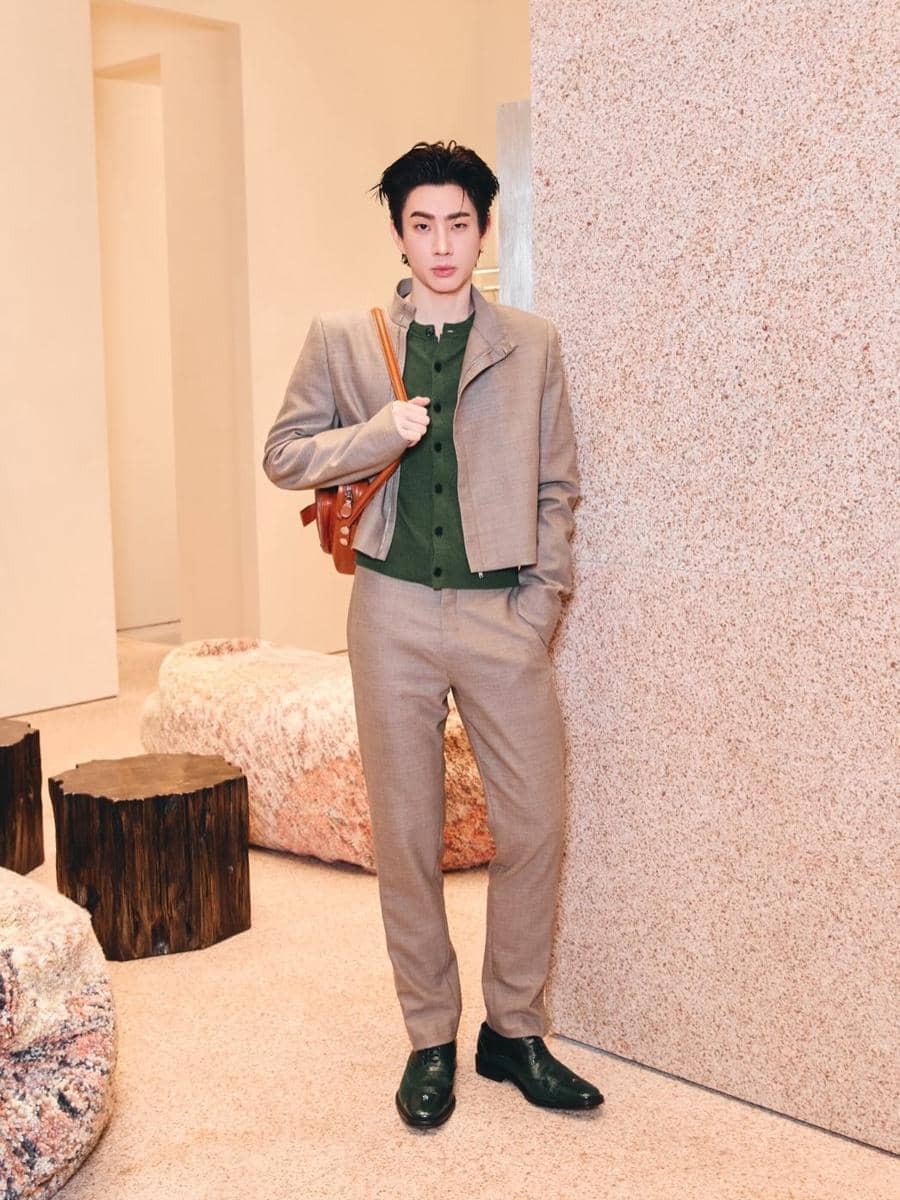 Seorang pria mengenakan setelan jas cokelat muda dengan kemeja hijau dan sepatu hijau gelap, berdiri bersandar di dinding dalam ruangan modern.