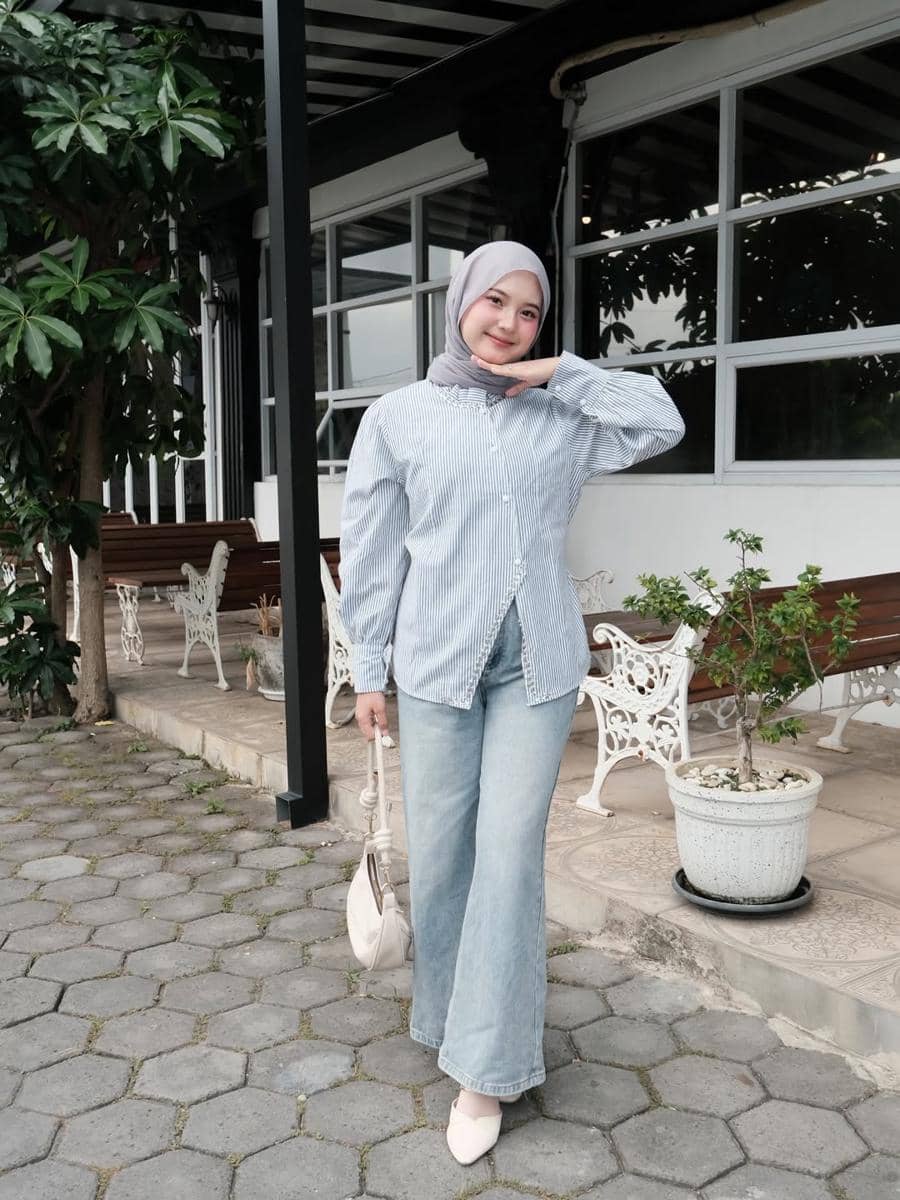 Seorang wanita berhijab berpose santai mengenakan kemeja bergaris biru dan celana jeans biru muda di area luar bangunan dengan kursi putih.