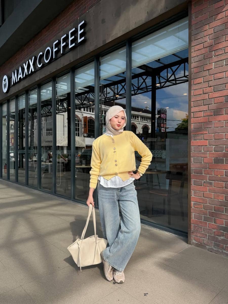 Seorang wanita berhijab mengenakan kardigan kuning dan celana jeans biru lebar berpose di depan kafe dengan dinding kaca dan bata merah.