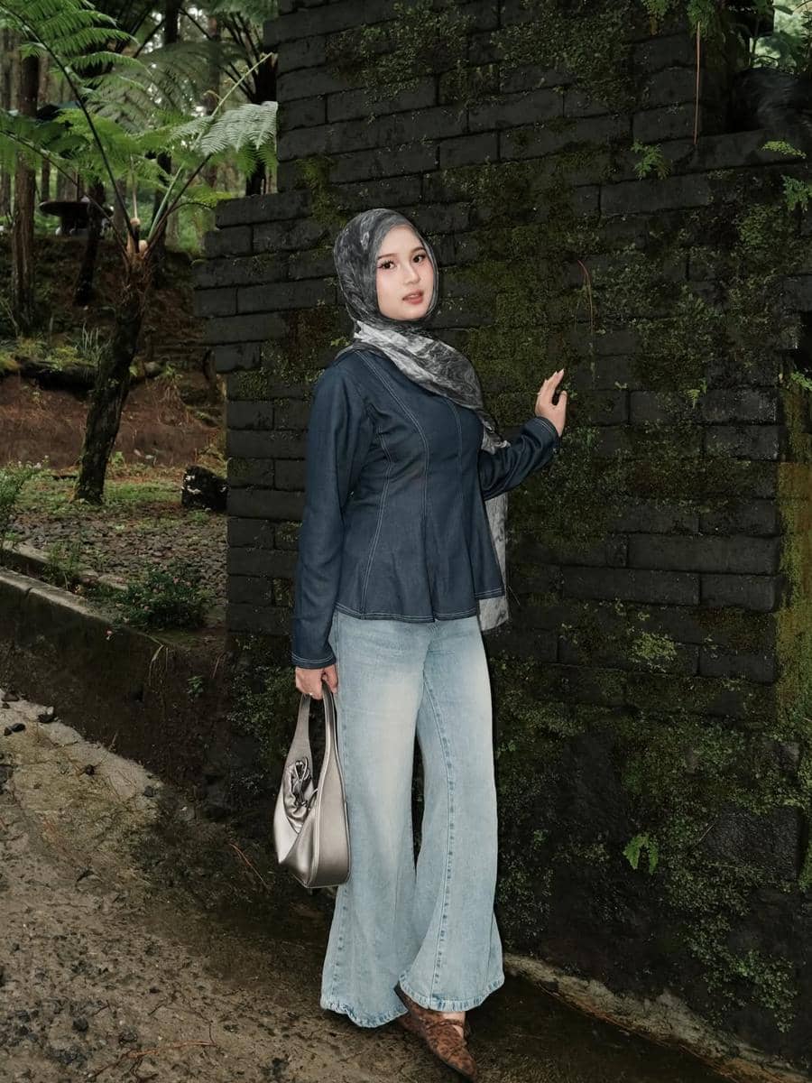 Seorang wanita berhijab mengenakan jaket denim biru dan celana jeans longgar berwarna terang berpose di dekat dinding batu berlumut di area hutan.