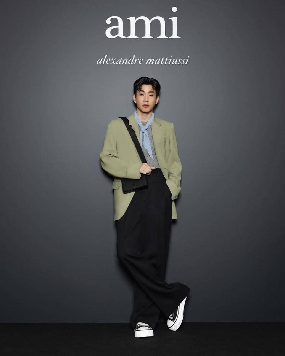 Seorang pria berpose di depan latar bertuliskan ami alexandre mattiussi mengenakan blazer hijau muda, celana hitam longgar, dan sepatu putih.