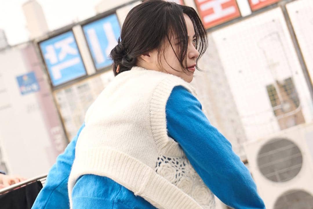 Seorang wanita berambut hitam mengenakan sweater biru dan rompi rajut putih sedang menoleh di area luar ruangan dengan latar papan bertulisan Korea.