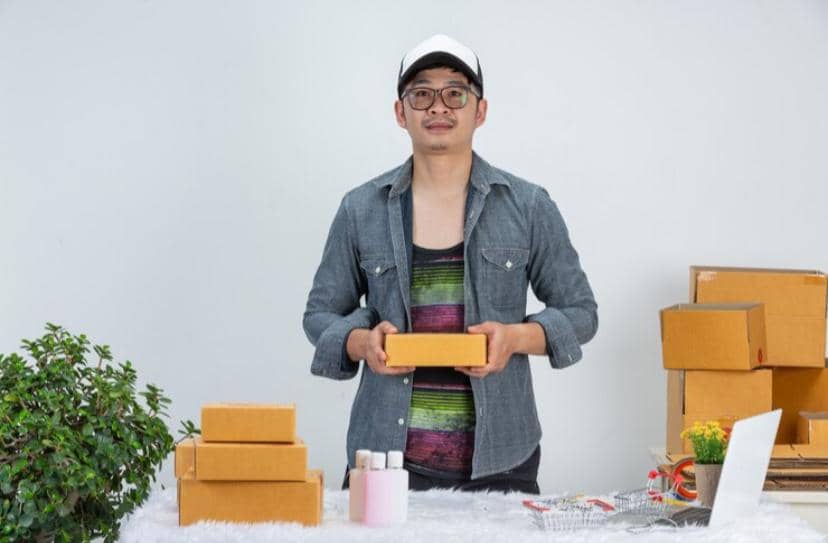 Seorang pria mengenakan topi dan kemeja denim memegang kotak paket di meja dengan beberapa kotak kardus dan tanaman hias di sekitarnya.