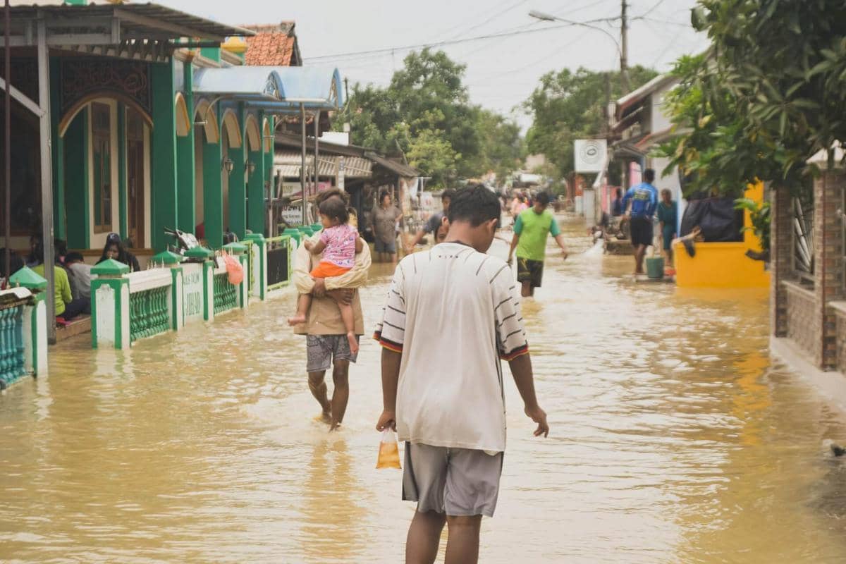 Warga berjalan di jalan perumahan yang tergenang banjir dengan air berwarna cokelat, beberapa membawa anak kecil di tengah genangan.